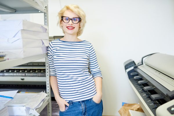 Portrait of blonde young woman standing by plotter machine and smiling at camera while posing in modern printing shop, copy space